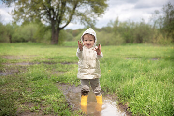 Cute child toddler in a warm vest and jacket stands in yellow rubber boots in a large puddle, Free children's walks in nature, mud and puddles