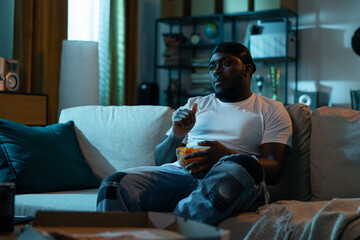 A man sits in a dark room at night lit by lights coming through a window from the street. Guy is watching television, soccer games, TV series while eating chips.