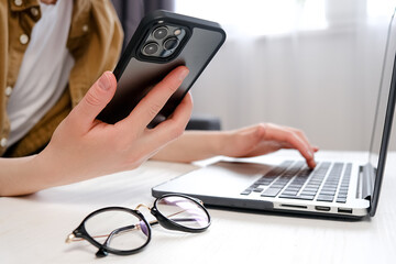 Fototapeta premium Close up of female hands holding smartphone and typing on computer keyboard, designer or student using laptop sitting on sofa at home, online learning, internet marketing, office workplace concept