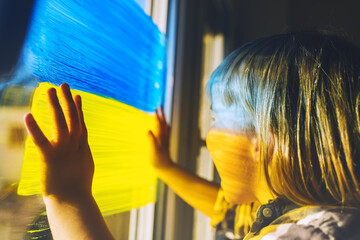 Fototapeta Little child with hands on yellow-blue flag image of Ukraine on window. Support Ukrainians. Concept, emotions. Color symbol image of flag of Ukraine on glass with paints. obraz