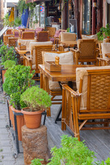 A row of massive wooden tables and wicker chairs with cushions in a cafe. Pots with flowers.