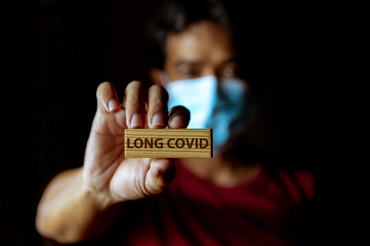 Masked Asian man show wooden sign with wording "Long Covid" in dark room.