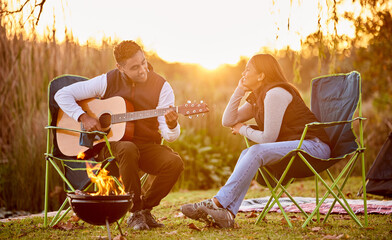 Life is better around the campfire. Shot of a you man playing his guitar to his wife outside while...