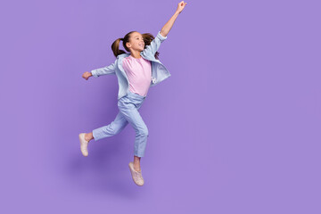 Fototapeta na wymiar Photo of cute excited little school girl wear denim shirt jumping high holding arm empty space isolated purple color background