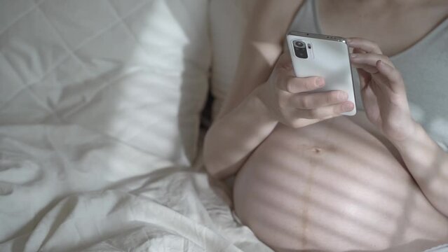 A pregnant woman uses the phone while relaxing in a home bed. Using a smartphone before the birth of a child. Women's health, remote consultation with a doctor, pregnancy period concept.