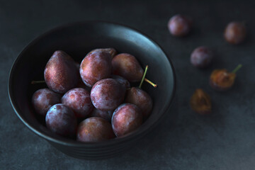Closeup Heap of fresh picked plums in a black bowl with dark blue grey background.