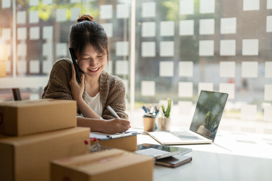 Young woman small business owner online shopping at home. confirm orders from customers with mobile phone preparing package product on background. SME entrepreneur or freelance concept