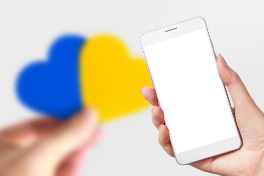 Ukrainian news channel. Female hand holds a mobile phone with a blank white screen and female hands hold a heart with a picture of the flag of Ukraine isolated over blue background. social networks