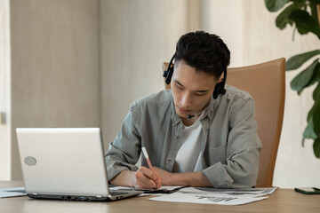 Asian assistant writes down payment details on paper helping customer to make purchase. Young man works using headset and laptop at table in office