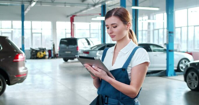Young Caucasian woman in blue overalls and white shirt slightly nods while using tablet in spacious repair shop. Portrait of female car mechanic at workplace.