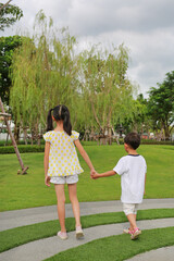 Little boy and girl child with hand in hands walking together in the garden. Back view of Sister and brother clasped in the summer park.