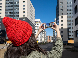 Female real estate agent in red winter hat taking a photo of property with her smartphone. Close-up of hands of realtor with the phone taking pictures of new residential buildings