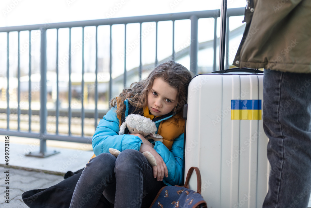 Wall mural Sad Ukrainian immigrant child with luggage waiting at train station, Ukrainian war concept.