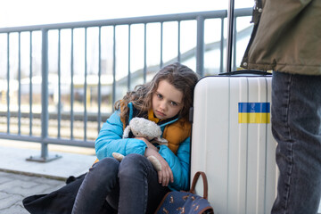 Sad Ukrainian immigrant child with luggage waiting at train station, Ukrainian war concept.