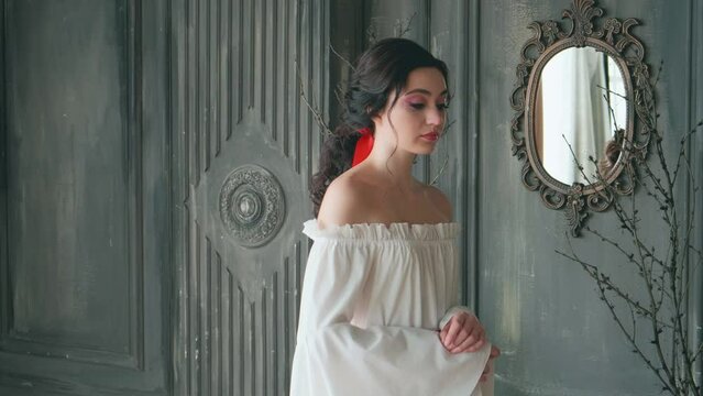Portrait sad young fairytale woman Snow White. Brunette wavy hair collected hairdo. White sexy vintage boho dress. Backdrop gothic style room medieval castle, fantasy girl princess. Old style mirror.