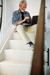 A senior man is typing on a laptop and sitting on the staircase inside.