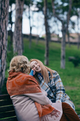 A young ginger woman is sitting with her grandmother and talking on the park bench in the cold...