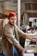 Vertical medium portrait of young Caucasian male carpenter wearing red knit cap working with drill press looking at camera