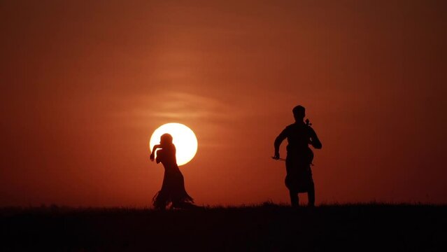 Silhouette of woman dancer and cellist opposite big sun in sky. Slow motion
