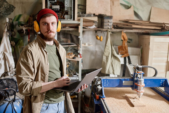 Horizontal medium portrait of young Caucasian man working in carpentry workshop holding laptop looking at camera