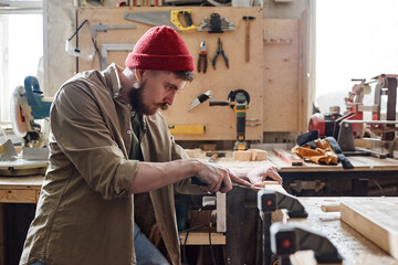 Side view of bearded Caucasian carpenter wearing red knit cap cutting mortise in wood plank using chisel
