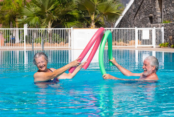 Cheerful adult happy senior couple having fun in outdoors swimming pool doing battle with swim noodles. Smiling people playing together in the pool enjoying good time and vacation