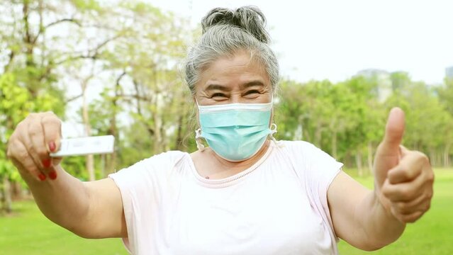 Elderly Asian woman wearing a medical mask tests with ATK KIT COVID-19 antigen test kit showing negative red line (negative: not infected) showing happy thumbs up.