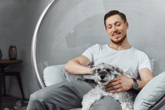 Young Man Sitting On A Chair In A Garden Play With A Gray Dog ​​sitting On Him. 