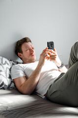 Caucasian attractive young man in white t-shirt woke up chatting, surfing, working on pc and making phone call on bed in bedroom interior. Modern device for remote communication