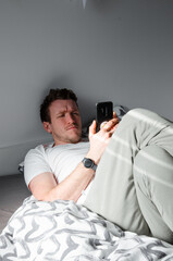 Caucasian attractive young man in white t-shirt woke up chatting, surfing, working on pc and making phone call on bed in bedroom interior. Modern device for remote communication