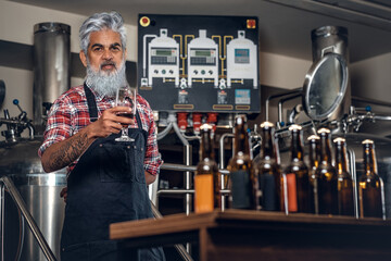 Production of quality beer. Old man brewer near table with beer looking at camera.