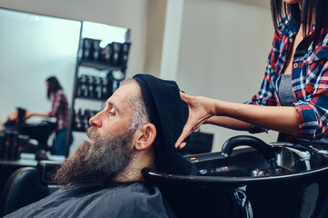 Shot of bearded elderly man hipster being washing by woman barber in hair salon in daytime.