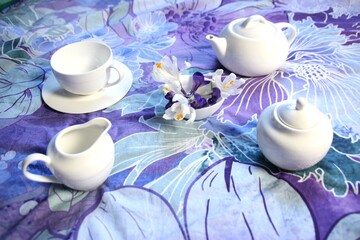 still life with crocus flowers and tea set of teapot, cup, sugar bowl, milk jug on colorful textile background