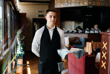 A young waiter in a stylish uniform stands with an exquisite dish on a tray near the table in a beautiful restaurant close-up. Restaurant activity, of the highest level.