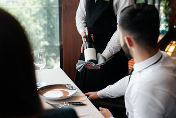 A young sommelier in a stylish apron demonstrates and offers fine wine to a beautiful couple in a restaurant. Customer service in an elite restaurant and a public restaurant. - obrazy, fototapety, plakaty