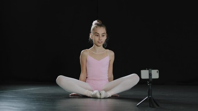 Teen girl dancer gymnast acrobat ballerina child making butterfly stretch exercise sitting on floor in dance studio hips stretching online video call remote distant class internet with phone on tripod