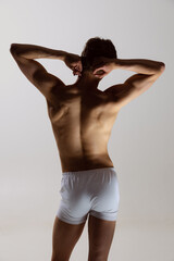 Fototapeta na wymiar Back view of young shirtless muscled man wearing white boxer-briefs standing isolated on gray background. Natural beauty of male body