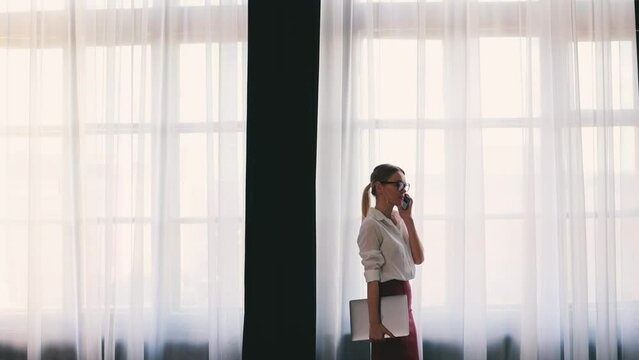 4k Elegant businesswoman in glasses and white shirt. Talking on the phone near big office window.