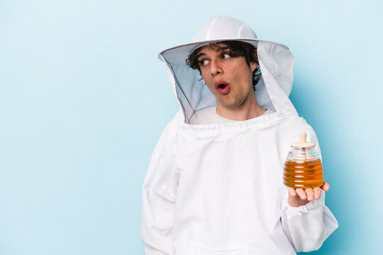 Young caucasian beekeeper man isolated on blue background is saying a secret hot braking news and looking aside