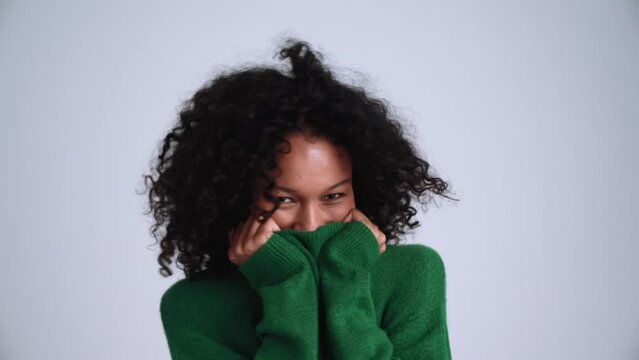 Smiling curly-haired woman pulling sweater over face in the grey studio