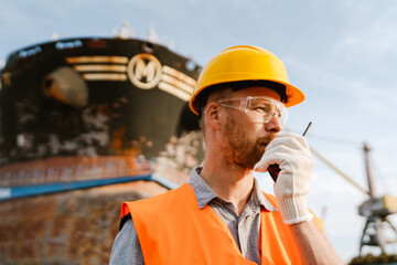 White man wearing helmet and vest using radio while working in port