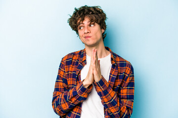 Young caucasian man isolated on blue background praying, showing devotion, religious person looking for divine inspiration.