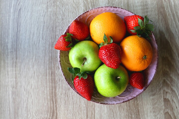 Pink bowl filled with fresh apples, oranges and strawberries on wooden table. Flat lay.