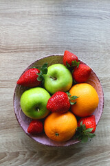 Pink bowl filled with fresh apples, oranges and strawberries on wooden table. Flat lay.