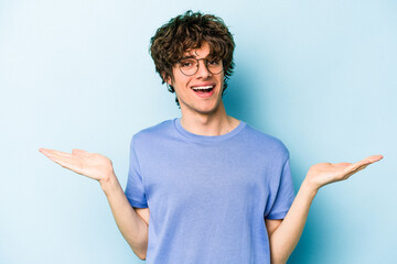 Young caucasian man isolated on blue background makes scale with arms, feels happy and confident.