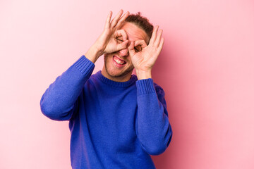 Young caucasian man isolated on pink background showing okay sign over eyes