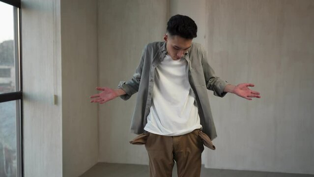 Asian man looks for money putting hands into inside-out pockets. Poor guy in casual clothes feels disappointed standing in empty room near big window