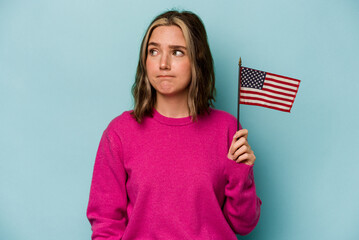 Young caucasian woman holding a American flag isolated on blue background confused, feels doubtful and unsure.