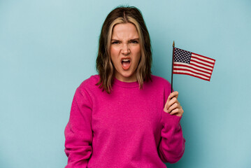 Young caucasian woman holding a American flag isolated on blue background screaming very angry and aggressive.