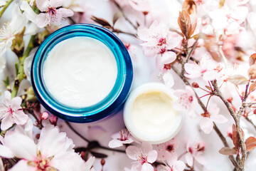 Obraz na płótnie Canvas Top view of cosmetic cream and concealer eyes creams with pink cherry flowers in a blue glass jar. Hygienic skincare lotion product.